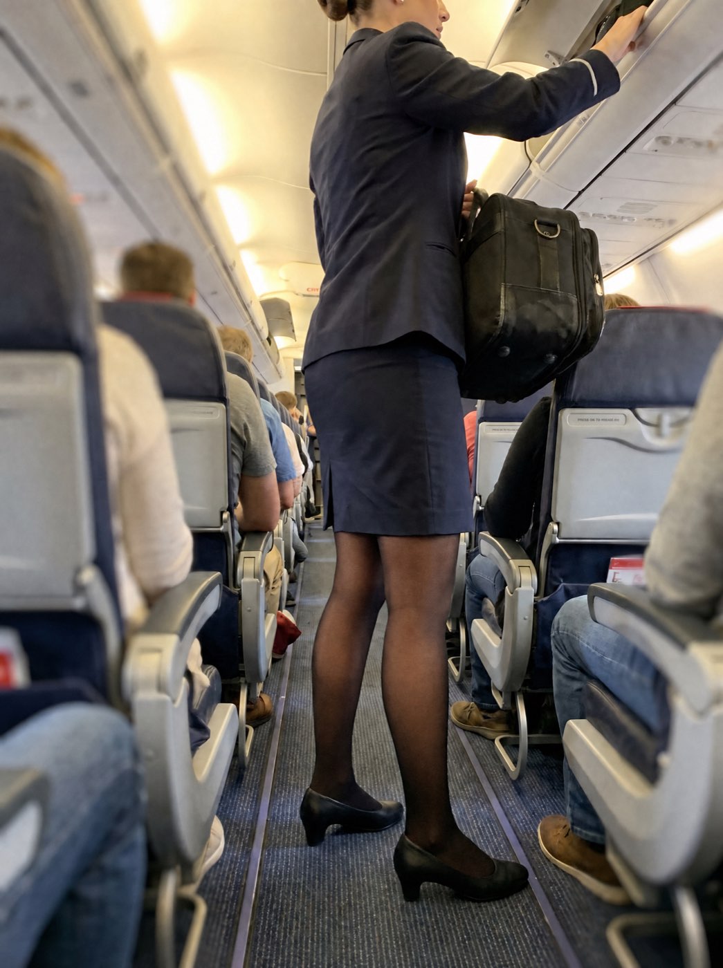 Flight attendant stowing a bag in the overhead bin, skin visible through sheer fleece-lined tights